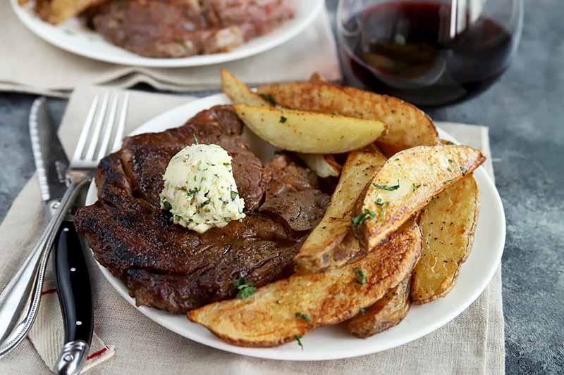 Horizontal image of a white plate with a seared large cut of beef topped with a dollop of herb butter next to potato wedges next to a glass of wine.