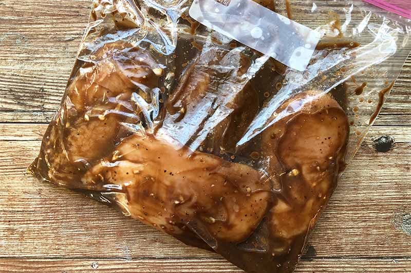 Horizontal image of a sealed plastic bag with raw poultry breasts and a dark brown marinade.