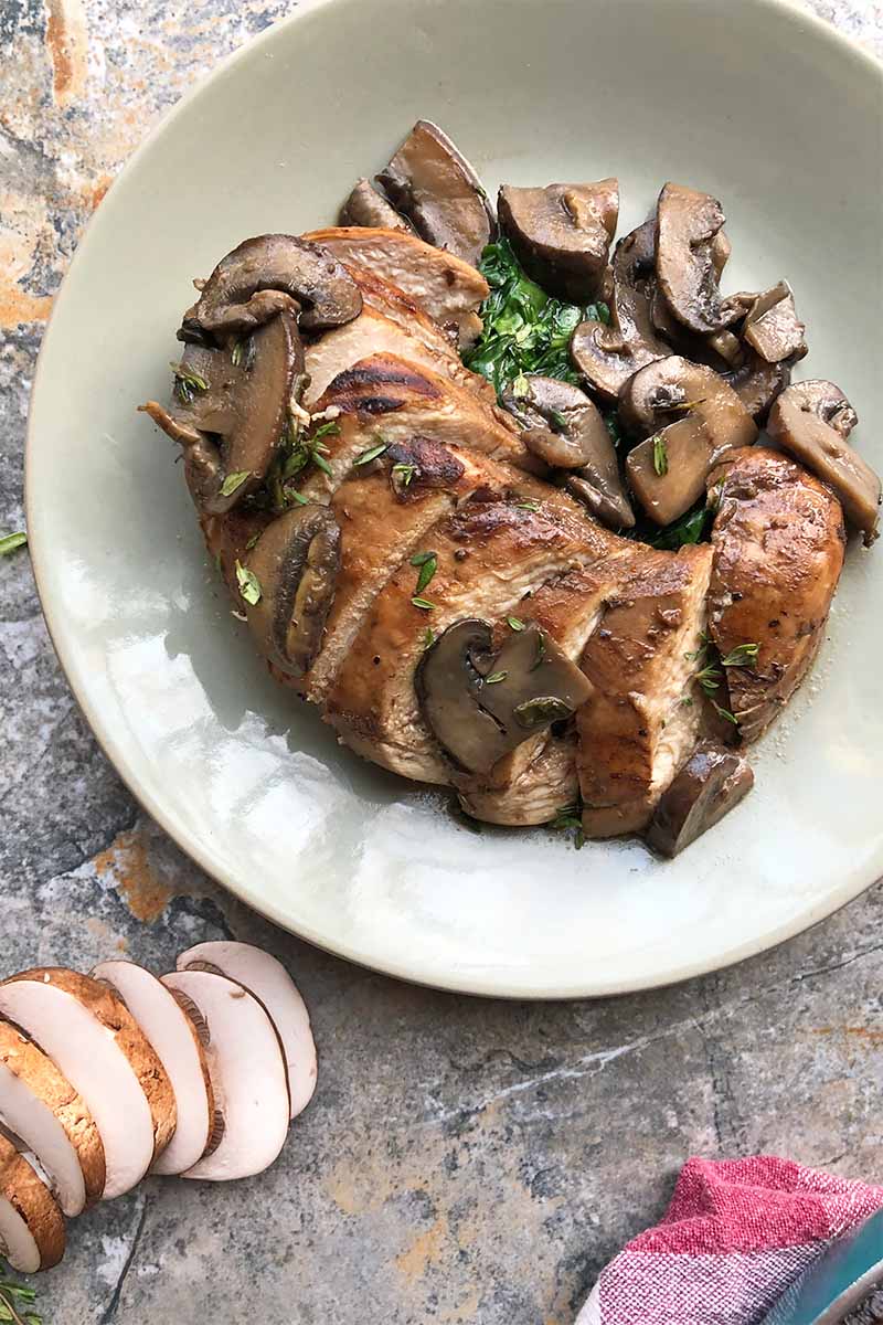 Vertical top-down image of sliced marinated chicken with mushrooms on a white plate on a gray slate surface.