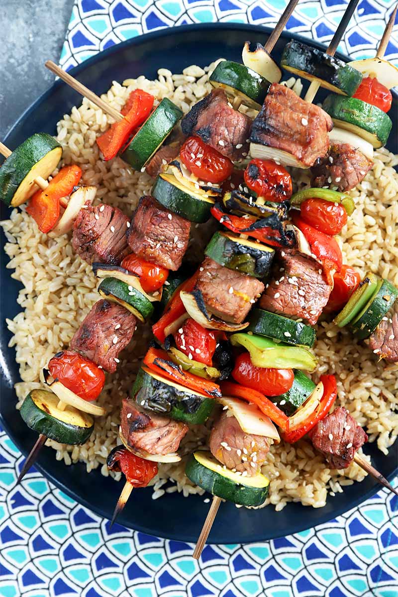 Vertical top-down image of cubes grilled meat and slices of vegetables on sticks over a bed of brown rice on a black plate.