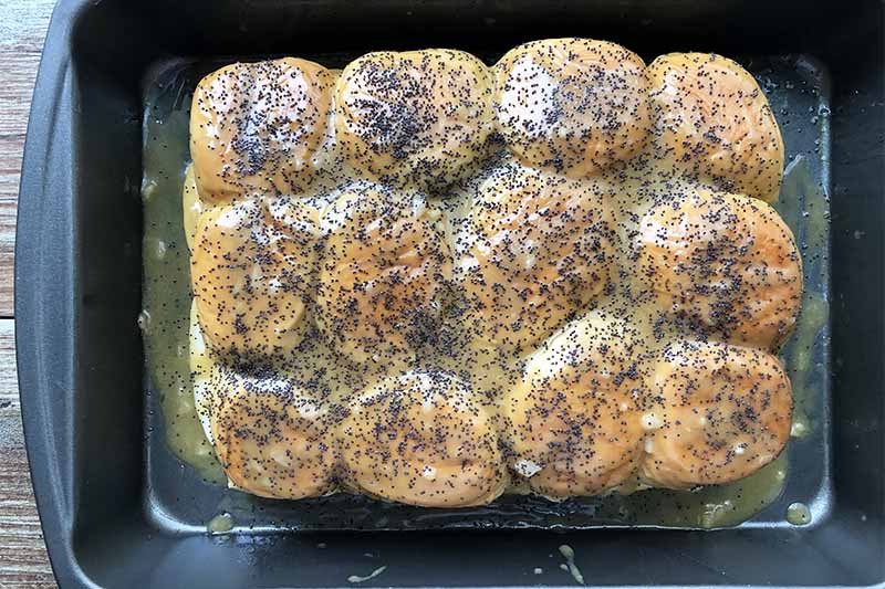 Horizontal top-down image of rows of buns covered in butter and poppyseeds in a pan.