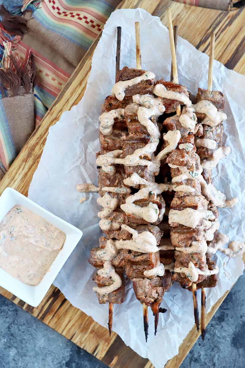 Vertical top-down image of a platter of grilled meat skewers drizzled with a yogurt sauce on top of parchment paper next to a white dish with more yogurt sauce.