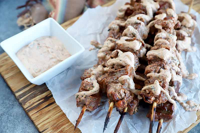 Horizontal image of a platter of grilled meat skewers drizzled with a yogurt sauce on top of parchment paper next to a white dish with more yogurt sauce.