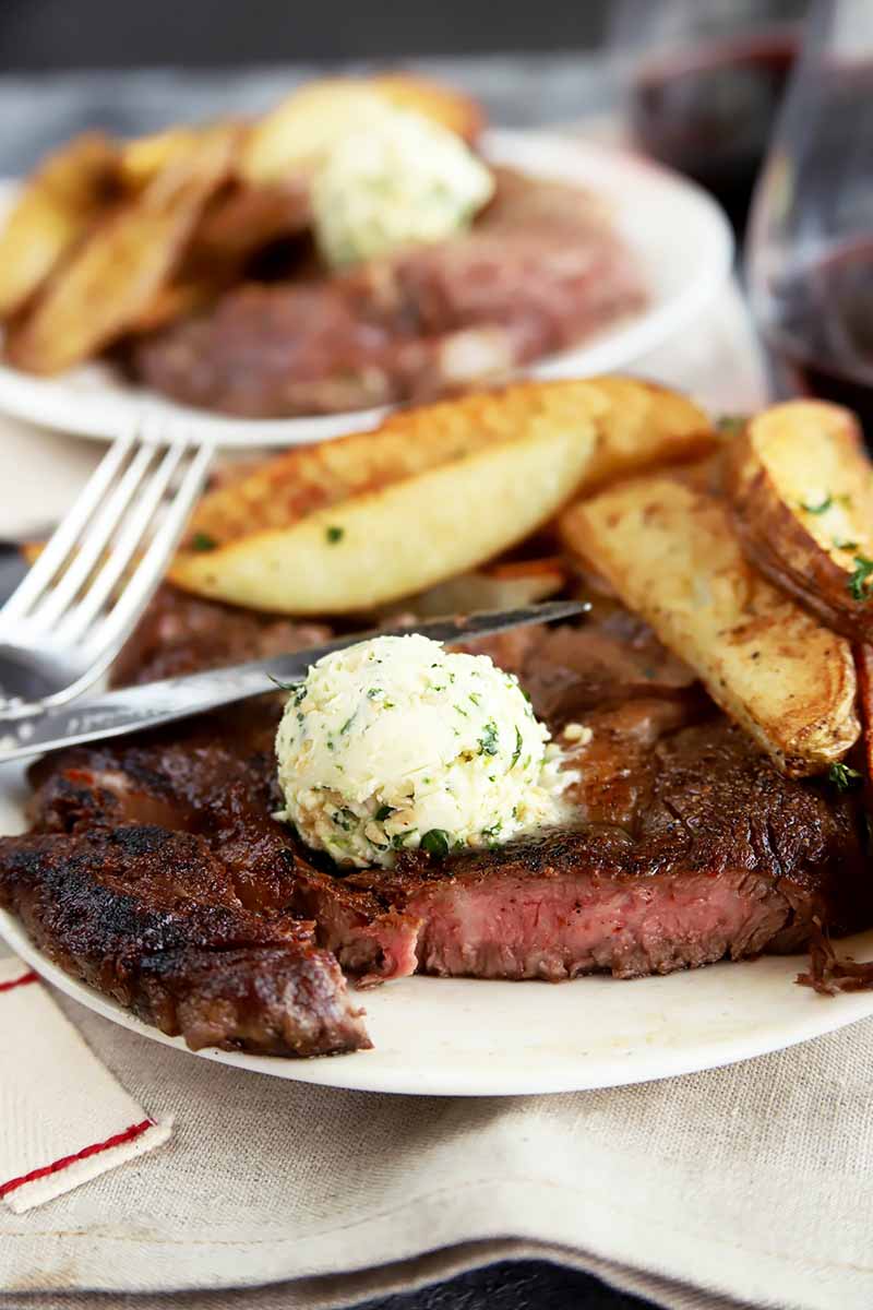 Vertical image of two plates of steak and potato wedges, with one steak cut into, next to silverware and glasses of red wine.