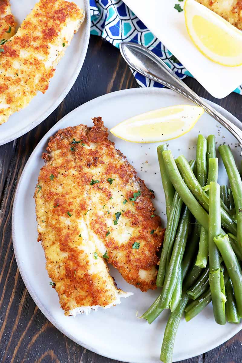 Vertical top-down image of plates with a cheese-encrusted tilapia next to lemon wedges, green beans, and a metal fork.