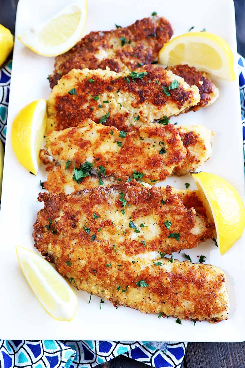 Vertical image of a white try with a row of cheese-encrusted fish surrounded by lemon wedges.