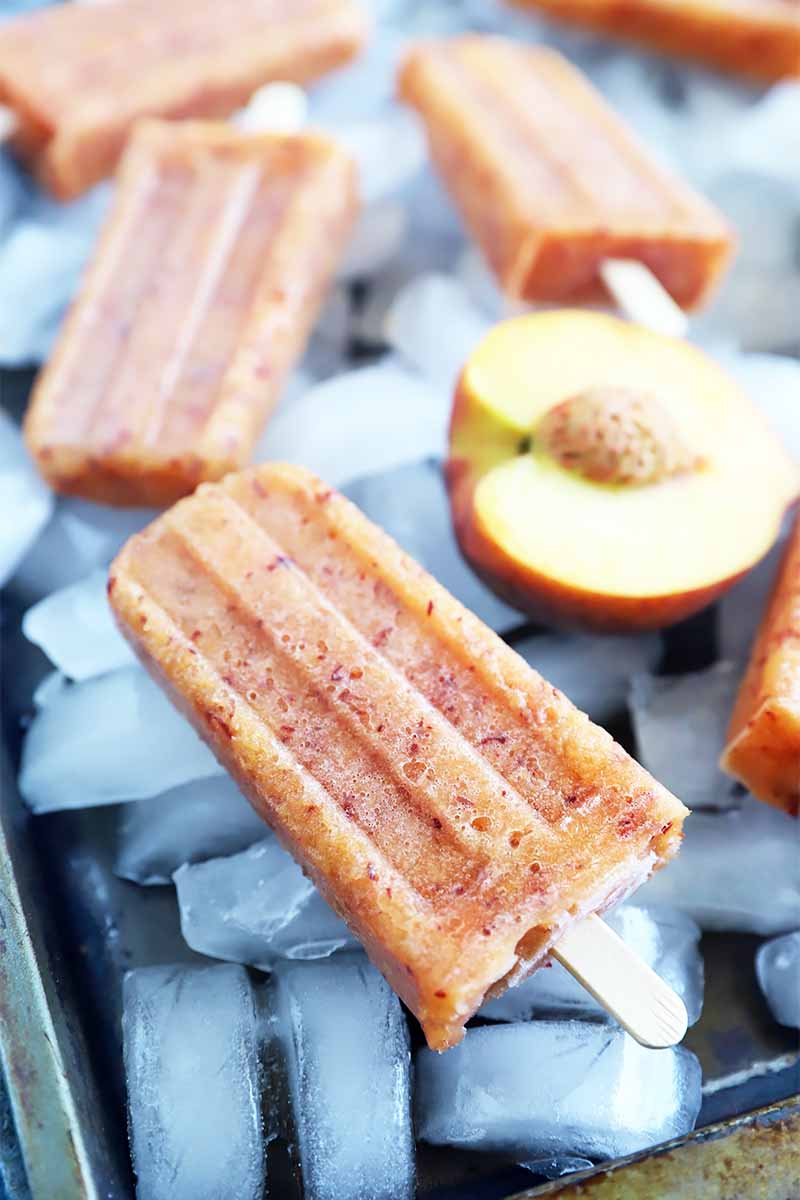 Vertical image of frozen light orange desserts inserted with wooden sticks on top of ice cubes next to half of a fruit.