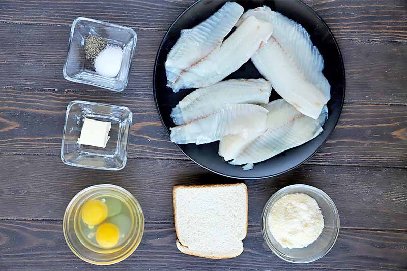 Horizontal image of a black plate with raw tilapia fillets, a slice of bread, and small glass dishes with assorted ingredients.