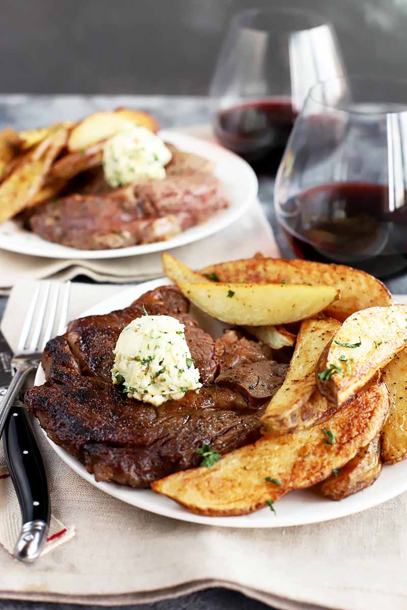 Vertical image of two white plate with a seared cut of meat topped with a dollop of butter next to potato wedges, with glasses of red wine next to the plates.