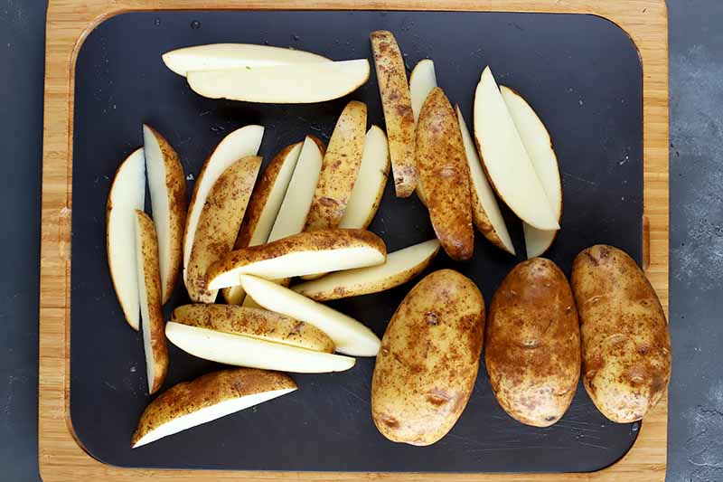 Horizontal image of whole potatoes and some cut in wedges on a black cutting board.