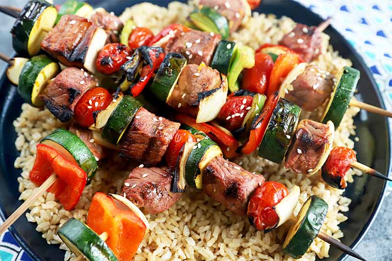 Horizontal image of pieces of beef and vegetables on sticks over brown rice on a black plate