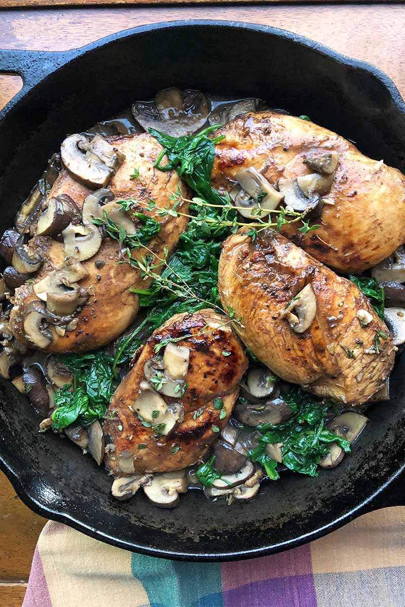 Vertical top-down image of a cast iron skillet with poultry, spinach, and mushrooms.