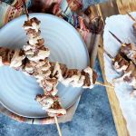 Horizontal image of two meat kebabs dried with yogurt sauce on a white plate, with more skewers next to them.