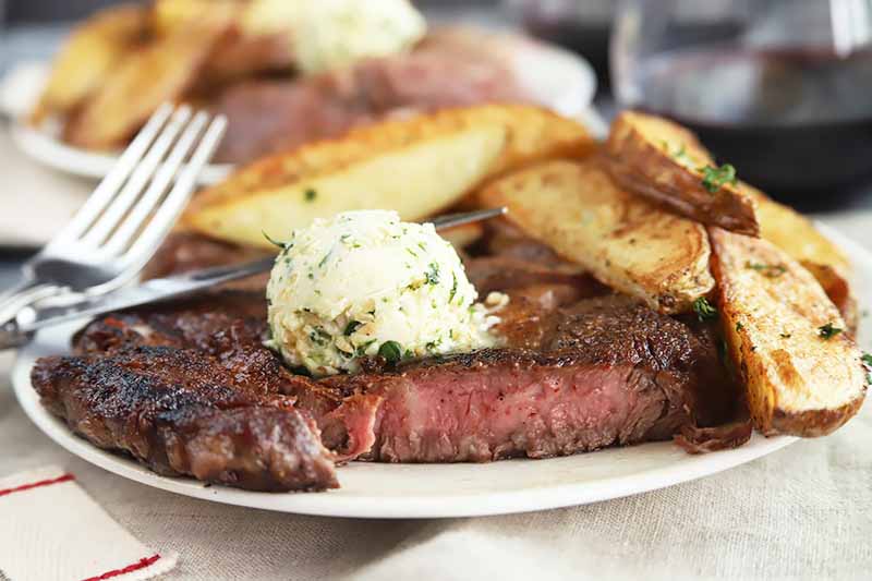Horizontal image of a white plate with a cut steak topped with herb butter with a side of potato wedges.