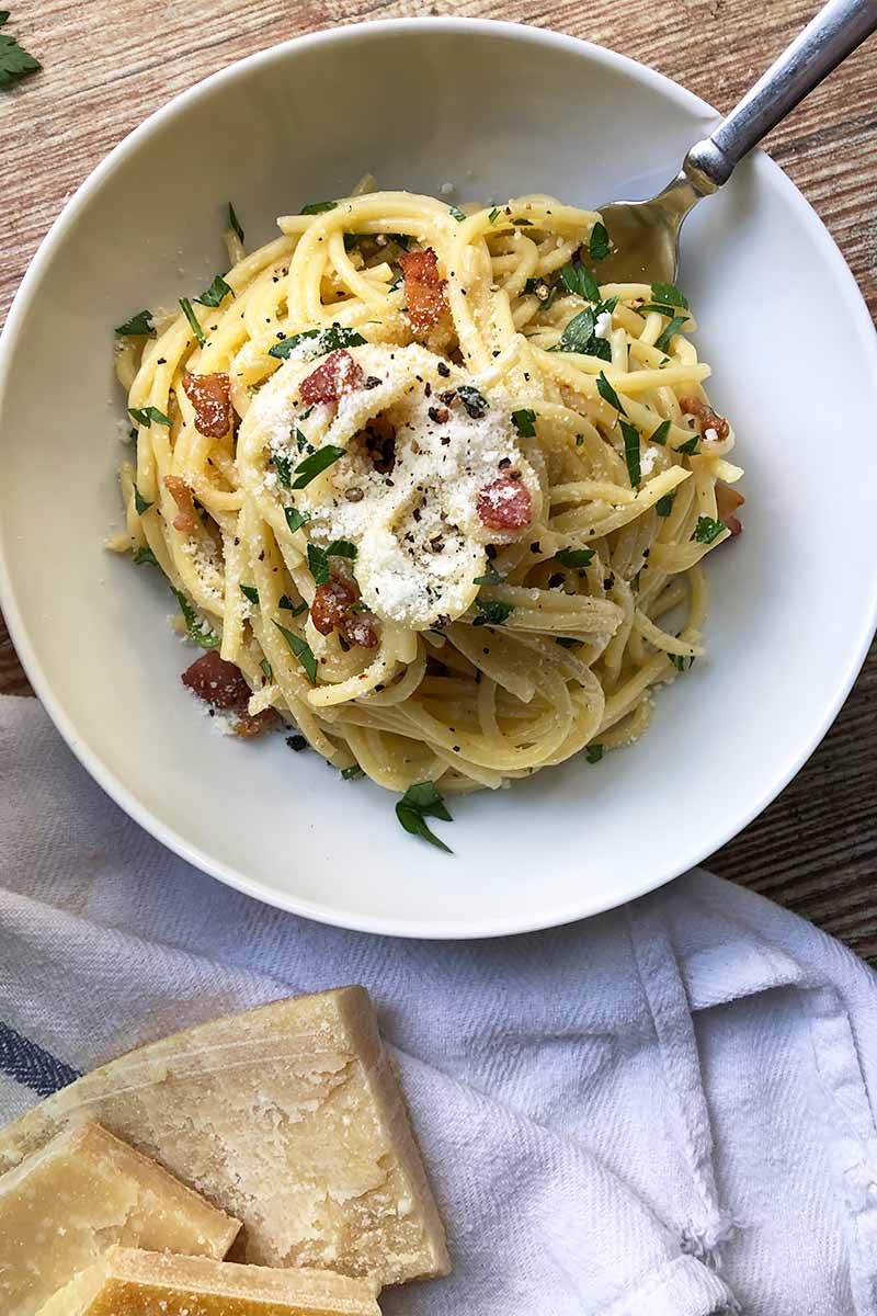 Vertical top-down image of spaghetti, herbs, bacon bits, and grated cheese in a white bowl on a white towel.