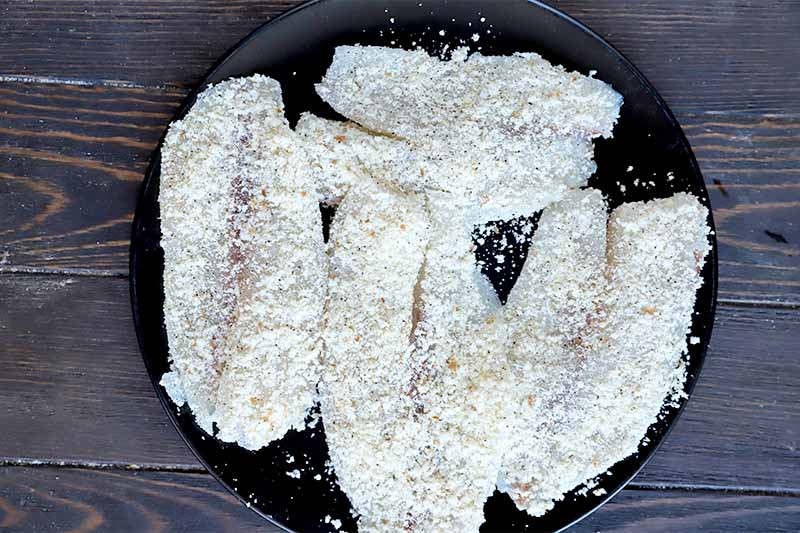 Horizontal image of uncooked and breaded fish fillets on a black plate on a dark wooden surface.