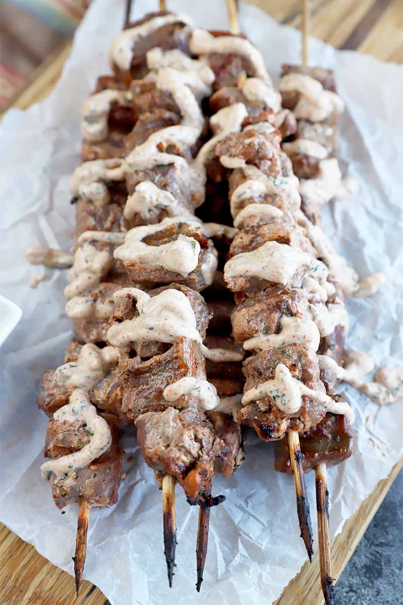 Vertical image of a stack of meat kebabs drizzled with a yogurt dressing on to of parchment paper.