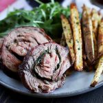 Horizontal image of a white plate with rolled beef medallions next to arugula and fries.