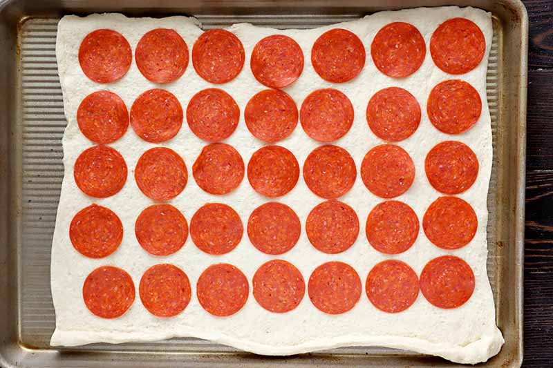 Horizontal image of a rectangular large piece of dough with clean, even rows of pepperoni slices on top.