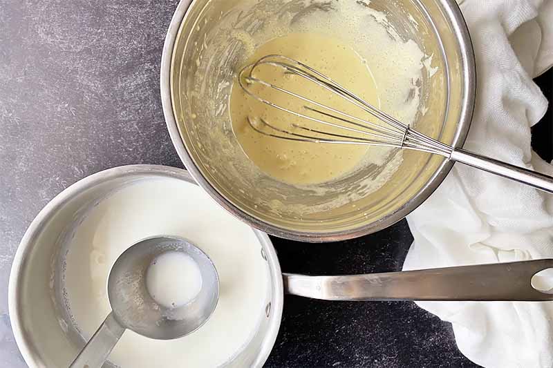 Horizontal image of a pot of milk with a ladle and a bowl of an egg mixture with a whisk.