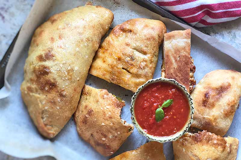 Horizontal image of calzones cut in assorted sizes on a baking sheet with a bowl of marinara.