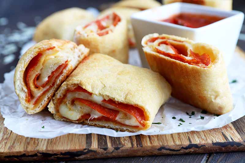 Horizontal image of four slices of pepperoni and cheese rolls on a baking sheet on a wooden cutting in front of marinara sauce.