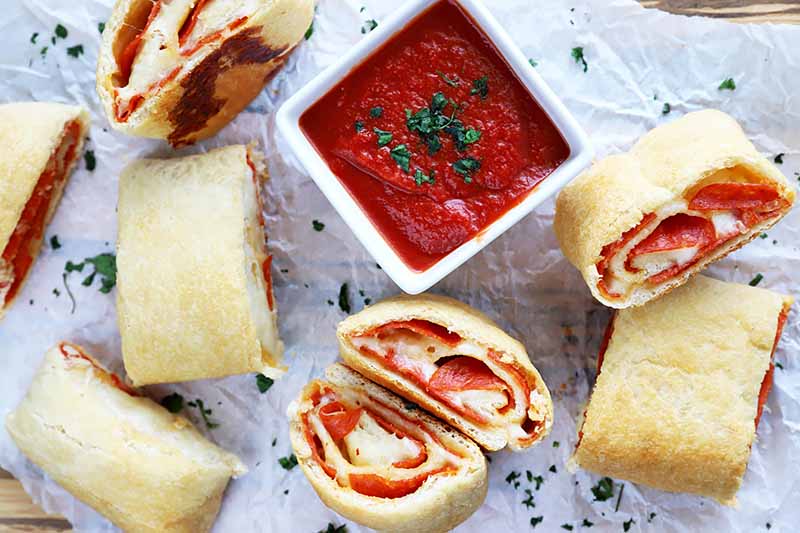 Horizontal image of slices of a meat and cheese roll on a baking sheet next to a white square bowl filled with marinara sauce.