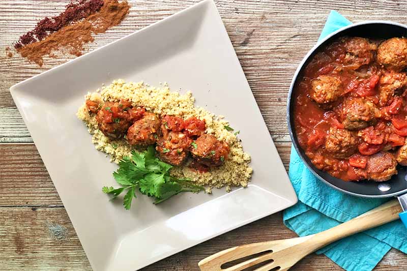 Horizontal image of a white square plate and a pan of meatballs in tomato sauce, with the ones on the plate over couscous and herbs.