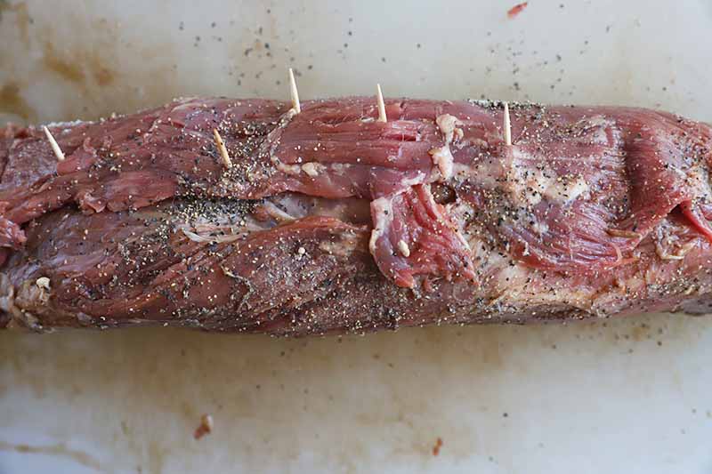 Horizontal image of a rolled log of raw beef held together by toothpicks on a cutting board.