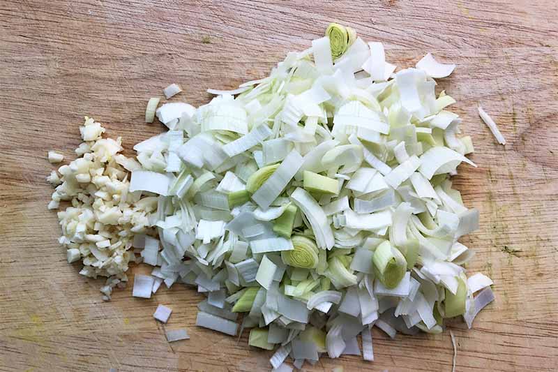 Horizontal image of chopped leeks and garlic on a wooden cutting board.