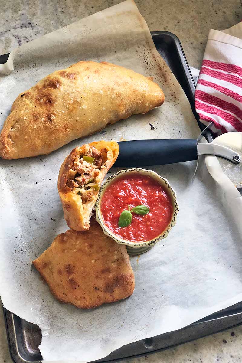 Vertical image of a whole calzone and one cut in half to reveal the meat filling on a baking sheet next to a slicer and a bowl of marinara.