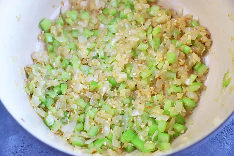 Horizontal image of finely chopped onion, garlic, and celery sweating in a large pot.