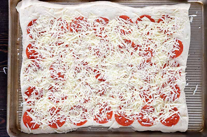 Horizontal image of a large rectangular piece of dough with rows of pepperoni slices and shredded cheese spread on top.
