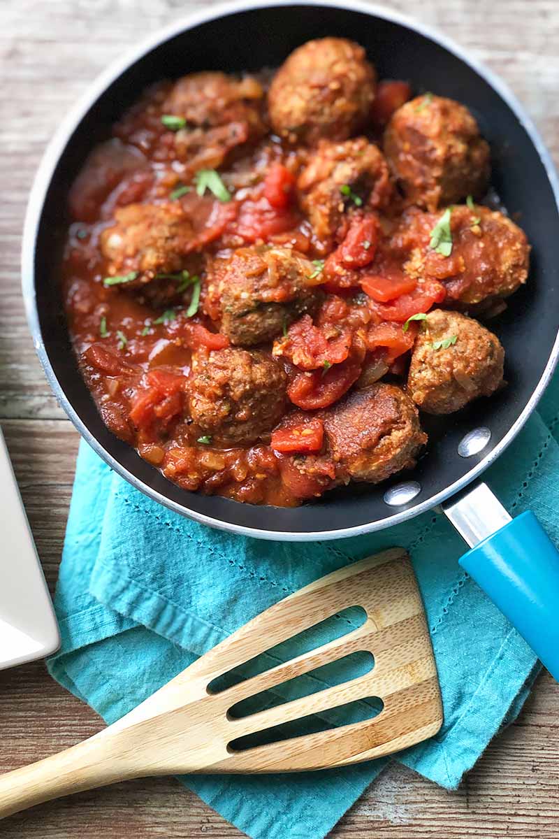 Vertical image of a pan filled with meatballs and stewed tomatoes on a blue napkin next to a wooden spoon.