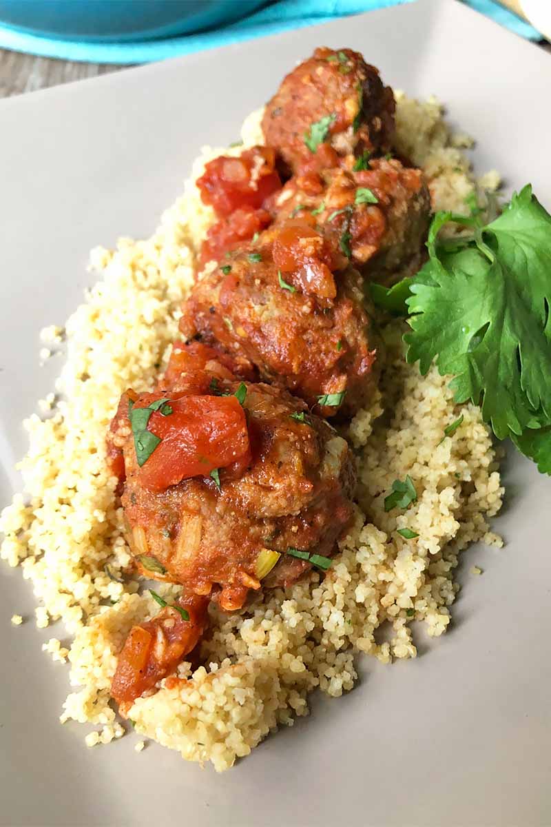 Vertical image of meatballs over couscous and fresh herbs on a white plate.