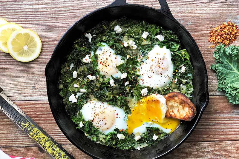 Horizontal image of a cast iron skillet filled with cooked greens and lightly cooked eggs next to lemon slices, red pepper flakes, and a zester.