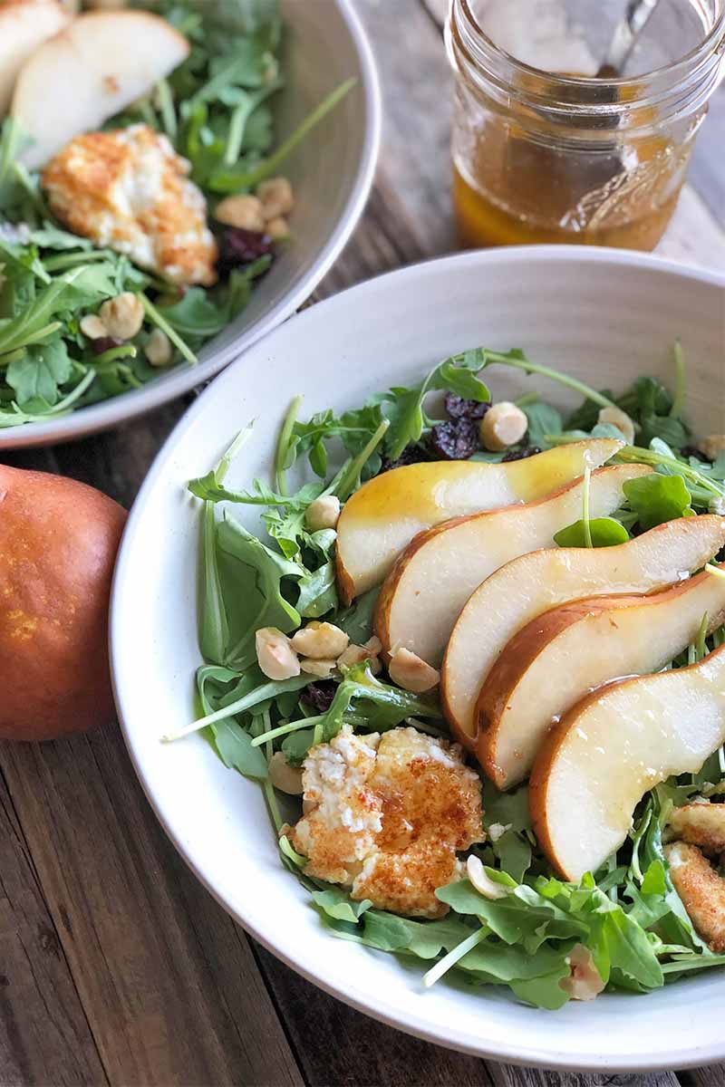 Vertical image of two white bowls with sliced pears and greens with fried chevre and chopped hazelnuts.