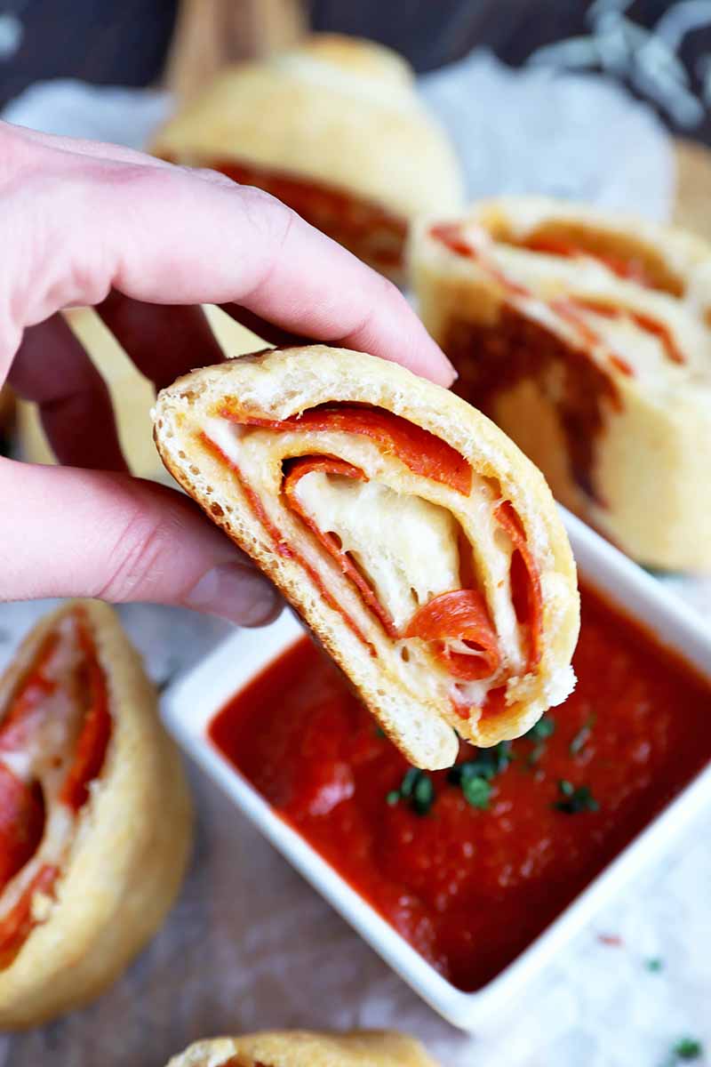 Vertical image of a hand dipping a meat and cheese roll in a white bowl filled with a marinara sauce.