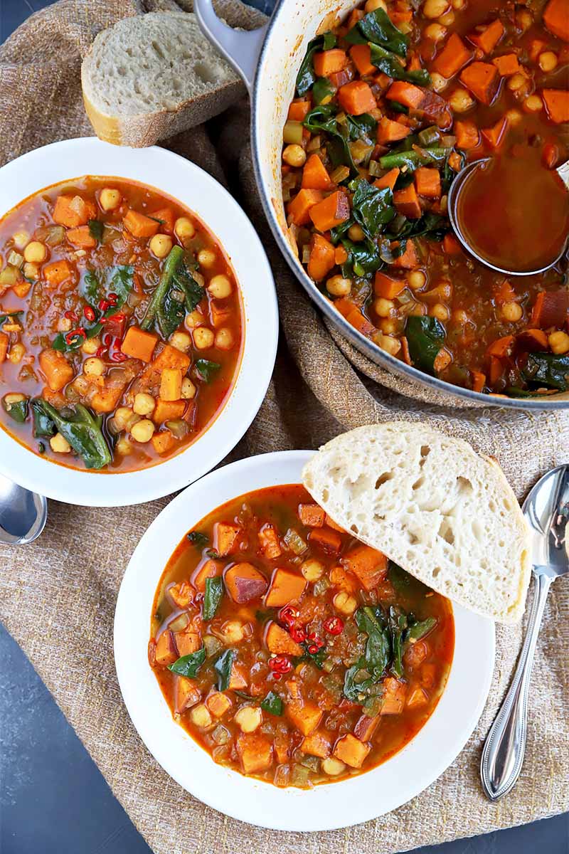 Vertical image of two bowls and a pot full of a hearty vegetarian stew on burlap.