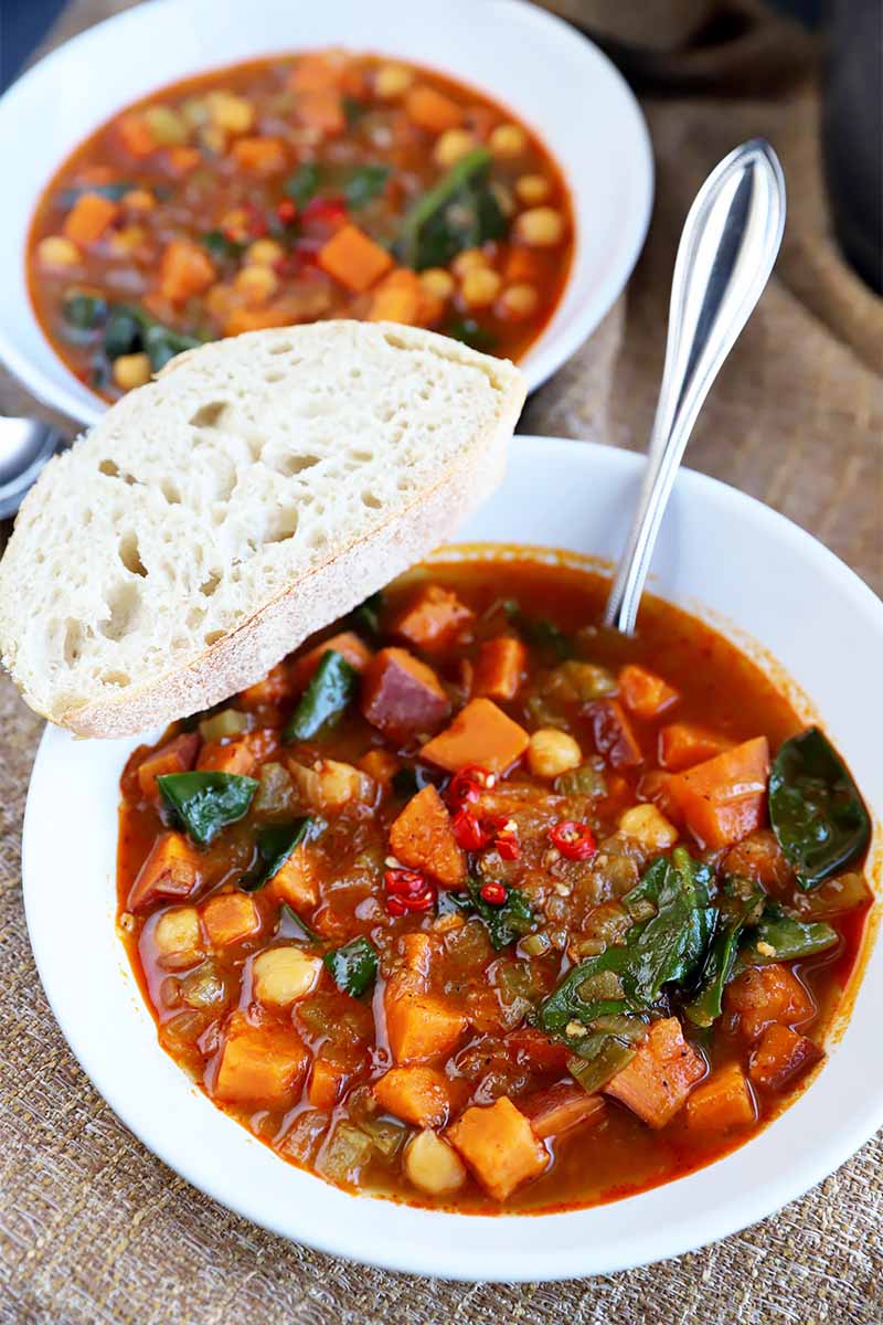 Vertical image of two white bowls with a vegetable stew in a tomato broth with a slice of bread and a metal spoon.