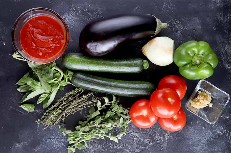 Horizontal image of assorted whole vegetables and herbs on a dark surface.