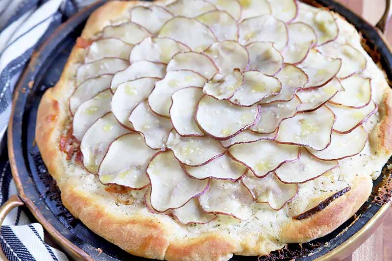 Horizontal image of a whole pizza topped with shingled thinly sliced potatoes on a pizza stone.