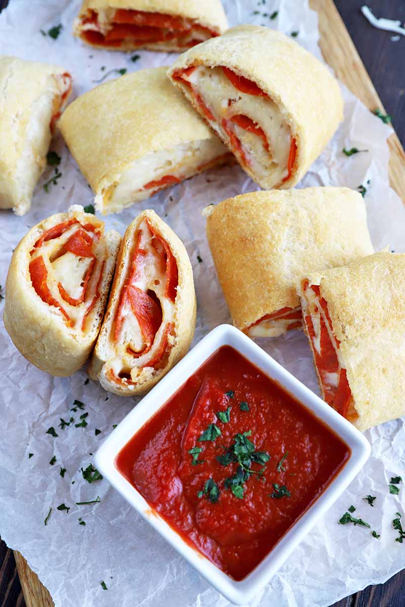 Vertical image of slices of a meat and cheese roll on a baking sheet next to a white square bowl filled with marinara sauce.
