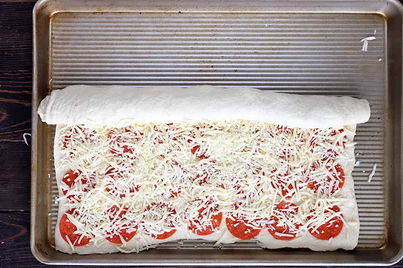 Horizontal image of rolling a long piece of dough with meat slices and shredded cheese in the center.