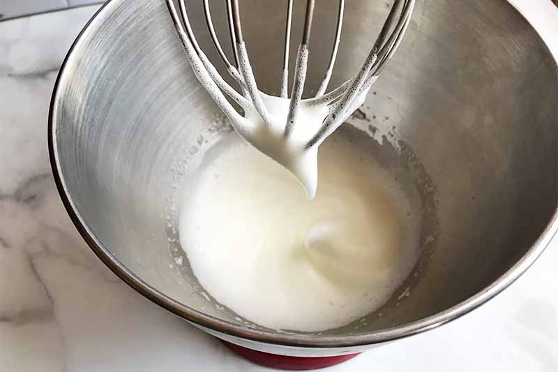 Horizontal image of a metal bowl with whipped egg whites, and a whisk with whipped egg whites on the tip.