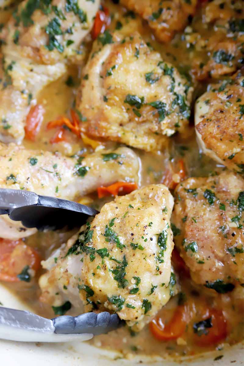 Vertical image of tongs holding up a piece of poultry in a pot with other pieces covered in an herb and white wine sauce with tomatoes.