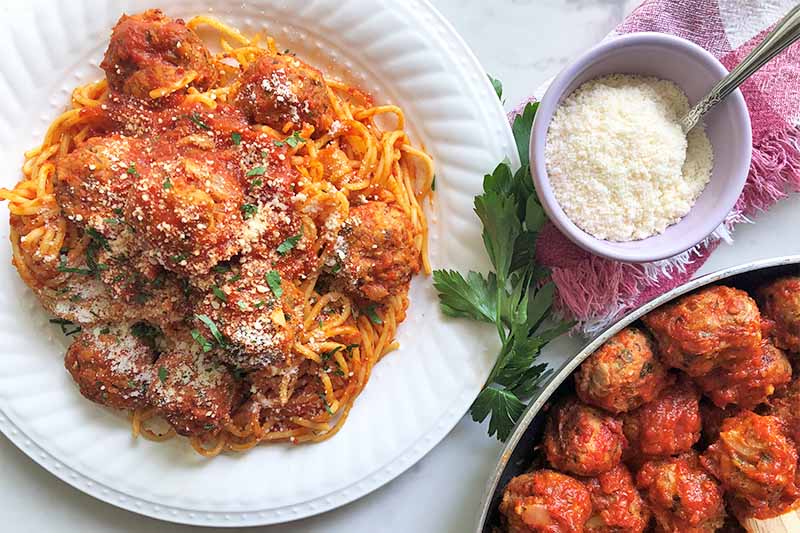 Horizontal image of a white plate with spaghetti and meatballs and marinara next to a pan of the same dish without the pasta and a bowl of grated cheese.