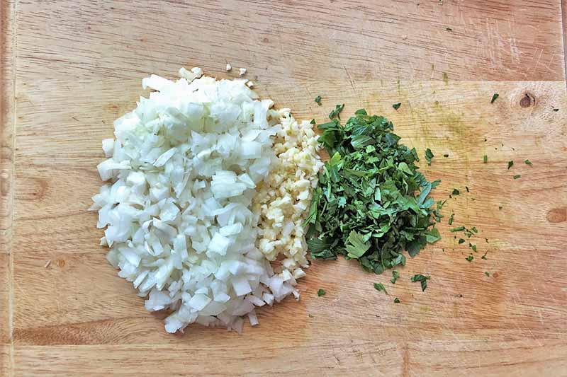 Horizontal image of a wooden cutting board with chopped onions, garlic, and fresh herbs.
