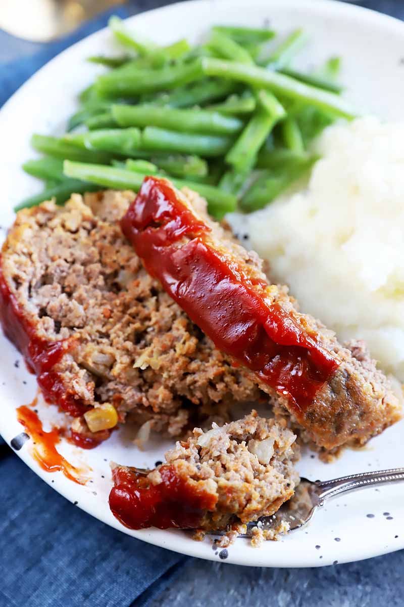 Vertical close-up image of a fork removing a large chunk from a beef dish with a ketchup glaze next to green beans and mashed potatoes on a white plate.