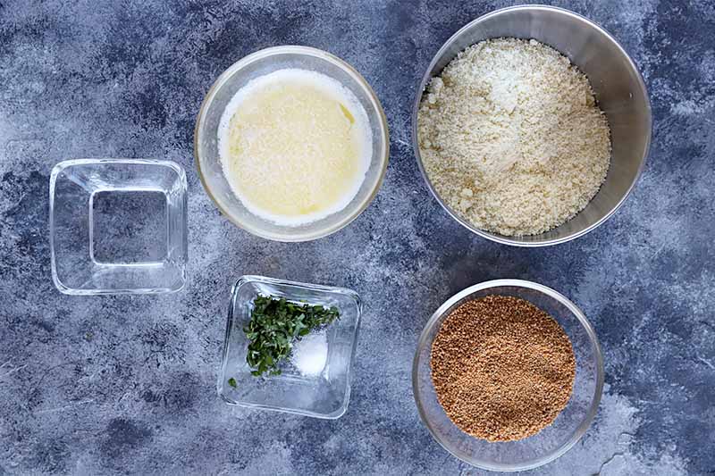 Horizontal image of bowls of almond meal, liquid ingredients, seasonings, and dark sugar on a gray surface.
